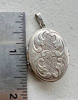 Close-up image of the front of the oval silver-tone locket next to a ruler showing size approximately 1.5 inches without bail.