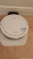 White circular Bissell robot vacuum sitting on white charging dock on a tiled floor. The vacuum has minimal visible dirt marks and Bissell logo on top.
