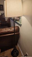 Full view of the floor lamp showing beige fabric shade and metal base next to brown couch and wall.