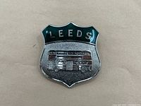 Front view of the shield-shaped badge with 'LEEDS' in green enamel and a raised double-decker bus image.