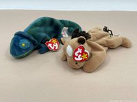 Two TY Beanie Babies plush toys together, one greenish-blue chameleon, the other tan dog (Chocolate). Both display TY heart tags.