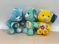 Three Care Bears plush toys shown together from the front, visible tags on two bears, different belly badges visible