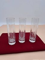 Front view of three tall crystal glasses standing upright on a red cloth, showing clear vertical fluted pattern and thick bases.