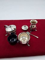 Photo of five silver plate items on a red surface showing the full set including covered sugar bowl, footed shaker, a small pot with spoon and blue glass insert.
