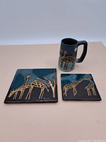 View of the mug with handle and two square tiles with giraffe motifs, showing overall style and color.