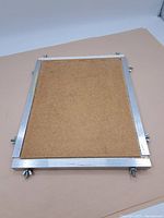 Rectangular metal painting frame laid flat showing backside with a brown backing board and silver metal frame with screws and wing nuts.