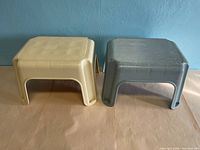 Two Rubbermaid stacking step stools, beige and gray, side by side on surface against blue wall.