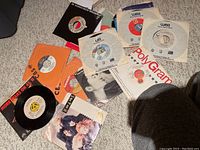 Multiple 45 rpm vinyl singles scattered on carpet including various paper sleeves and picture sleeves.