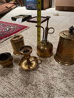 Five assorted brass items arranged on carpet with measuring tape for size reference; candlestick holder, ashtray, sprayer, covered mug, and small bowl.