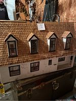 Front view of a large vintage wooden dollhouse with worn white painted walls, brown shingle roof with dormer windows, and black framed windows.