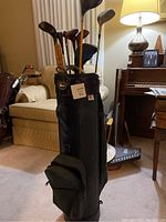 Black golf bag standing upright with visible pockets and golf clubs inside near a couch in a living room setting.