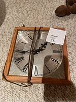 Front of handmade square wall clock showing wooden frame, metal face with Roman numerals and black clock hands wrapped with cord.