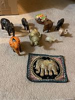Nine small elephant figurines in different materials and colors arranged on carpet along with embroidered textile piece