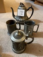 Photo showing the full set: large pewter pitcher with curved handle and lid, and three pewter steins/mugs including one with ornate relief.
