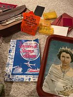 Shows assortment of collectible stamps in boxes, orange bag of world-wide stamps, Universal Stamp Album, metal Queen Elizabeth II tray, and other small containers on carpet.