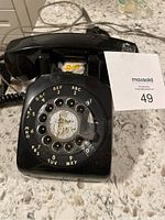 Front view of a black rotary dial telephone showing the rotary dial and base with some sticker residue and wear.