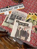 Photo showing commemorative newspapers titled 'The Kingston Hip Standard' and a Kingston Frontenacs road sign 'Tragically Hip Way' placed on patterned tablecloth.