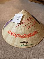 Photo showing front and side view of natural woven straw Vietnamese conical hat with embroidered red text 'SÀI GÒN DU LỊCH', red bridge, and black and green boat design.
