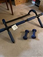 Full view of black chin up bar with padded blue grips, laying on carpet, alongside two blue dumbbells.