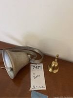 Small golden hand bell and larger hanging metal bell shown on wooden surface against white wall with auction tag.