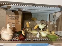 Front view of shelf with assorted home decor items including ceramic duck gravy boat, wooden duck figure, glass dishes, candle holders, and flatware caddy box.