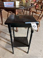 Small black wooden table with four turned legs and a lower shelf, showing surface scratches and some wear in a basement storage-like setting.