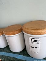 Side view of three white ceramic canisters with round wooden lids, showing size variation and a label.