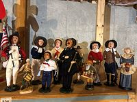 Full view of complete set of 11 Byers Choice holiday carolers on wooden shelf in basement setting