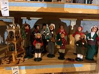 Group of Byers Choice Christmas Caroler figurines on shelf, various winter attire and holding miniature Christmas decorations, wooden village display parts in background.