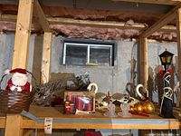 Photo of assorted Christmas decoration items on wooden shelving in basement including Santa figurine, swan candle holders, candle set, gold ornaments, lantern, and welcome mat.