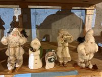 Five Christmas-themed figurines including Santas, snowman, and angel arranged on a wooden shelf in a basement setting.