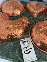 Set of five copper kitchen molds arranged on countertop showing varying shapes and embossed designs including a rooster, heart and floral motifs.