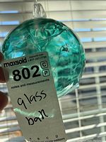 Photo showing the turquoise blown glass friendship ball held up against window blinds for lighting, showing internal swirled glass patterns and the hanging loop at the top.