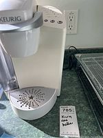White Keurig coffee maker on countertop with drip tray and power cord visible.