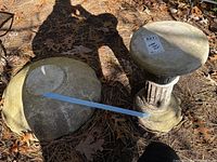 Two stone decorative pieces outdoors on ground with dry leaves, one is a pedestal birdbath and the other is a rounded stone piece.
