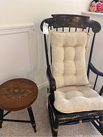Front view of black vintage rocking chair with beige cushion and small decorative wooden stool beside it.