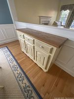 Angled view of buffet side table showing wooden top and painted cream base with decorative motifs.