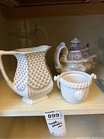 Photo showing three ceramic items on shelf: tall pitcher with patterned body, basketweave sugar bowl, and ornate purple floral teapot.
