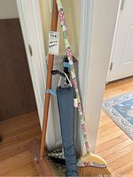 Three brooms, floral dustpan, and cane clustered in a corner indoors on hardwood floor.