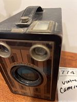 Front and side angle of vintage box camera showing the lens, two circular viewfinder windows, and leather strap on top.