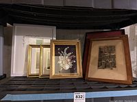 Wide view of multiple framed artworks and pictures on a shelf including floral painting, vintage photo, gold frames, and white frames