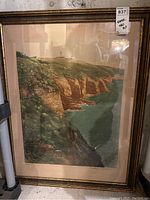 Framed coastal wall art with lighthouse on grassy cliffs over blue-green water, gold frame, beige matting.