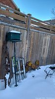 Two green metal bird feeders mounted on poles against a wooden fence with snow on ground. Several metal sunflower stakes and garden art pieces visible.