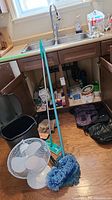 Photo showing a kitchen area with cleaning supplies beneath the sink including a black step waste can, white oscillating table fan, blue feather dusters, and mop handles.