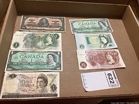 Photo showing seven different paper currency bills from Commonwealth countries placed in a cardboard box, featuring various denominations and portraits of historical figures such as Queen Elizabeth II and King George VI.