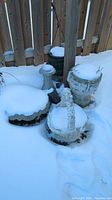 Four concrete garden items covered partly in snow including a bird bath, basket, and planters.