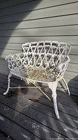 Front angle view of white painted iron garden bench showing decorative scroll backrest, seat lattice and armrest detailing. Bench surface shows weathering and rust spots.