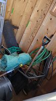 Photo of assorted gardening tools including green coiled hose, moving dolly and part of a weed eater inside a wooden shed