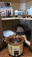 Photo showing Black & Decker toaster box, electric kettle, enameled roasting pan, white sandwich press, copper Roman pot, and crockpot with glass lid and electrical cord inside a cabinet.