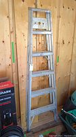 Full view of the aluminum A-frame ladder standing against a wooden wall, showing size and condition.